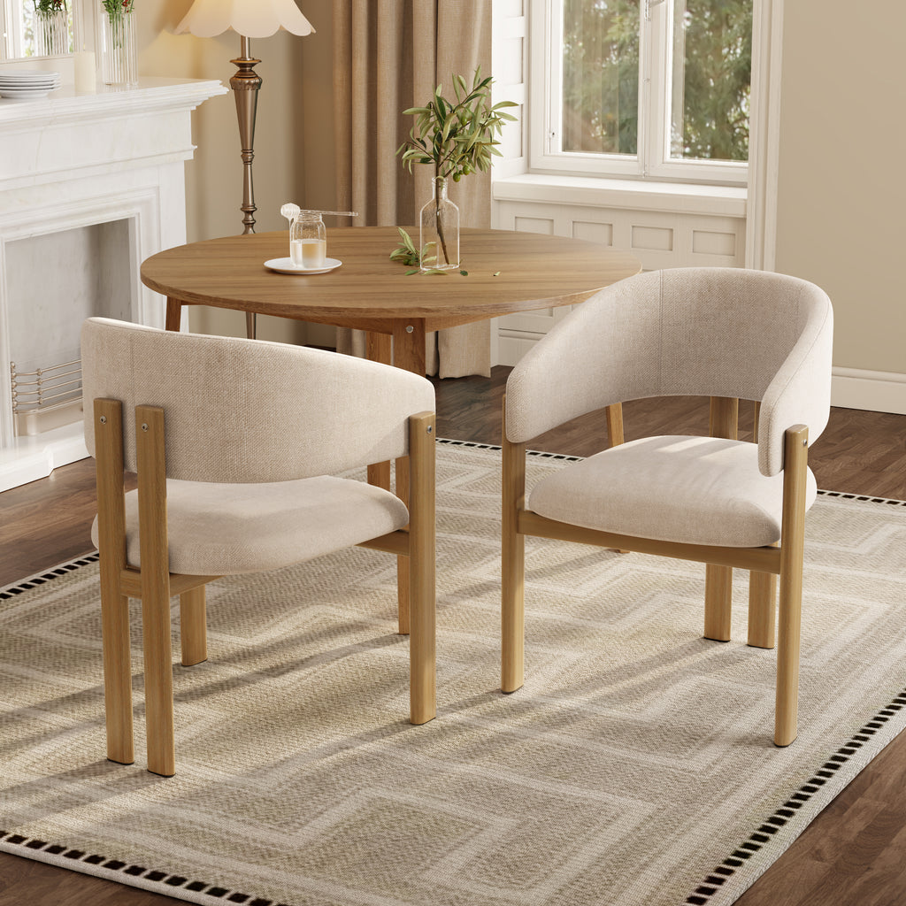 Two upholstered dining chairs with light wood frames arranged around a round wooden table in a bright, modern dining room with a patterned rug, fireplace, and window.