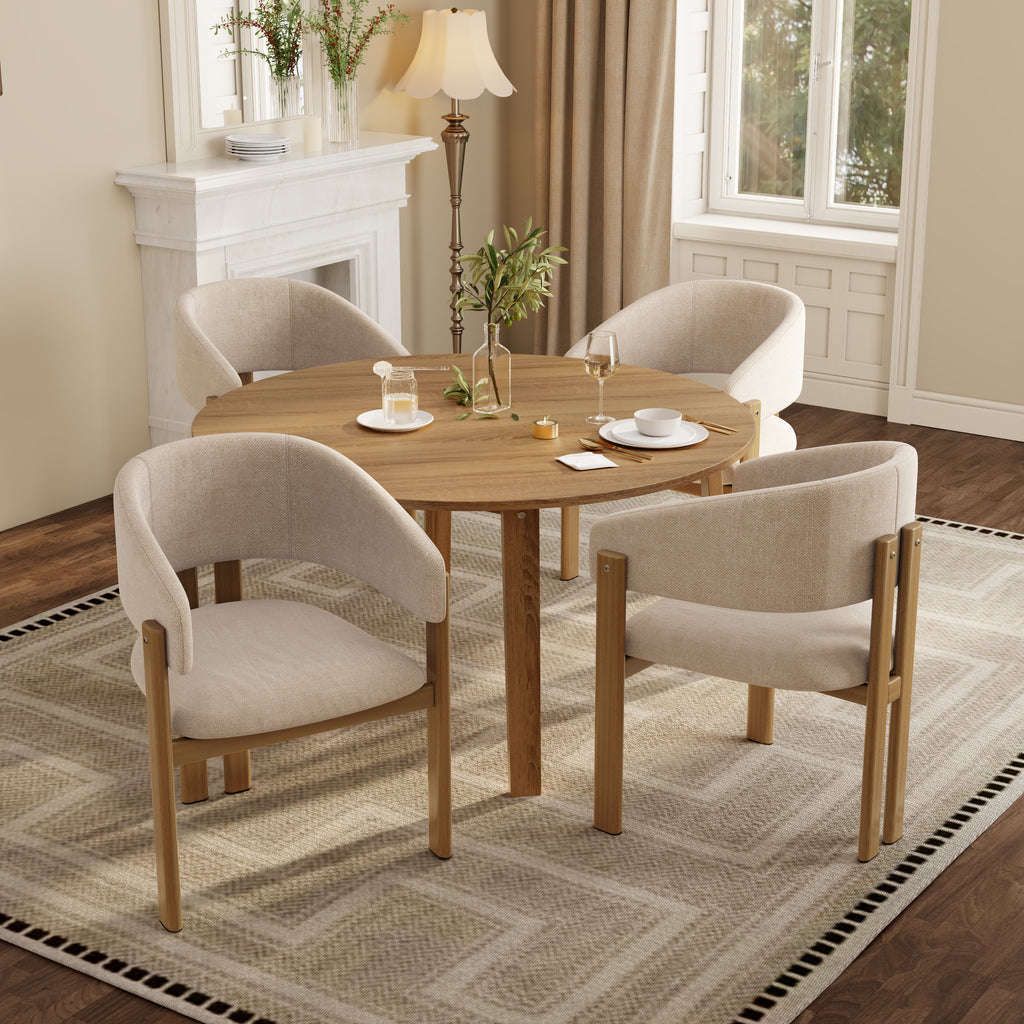 A round wooden dining table with four upholstered armchairs in a warm beige fabric, styled in a bright, modern dining room with a fireplace, lamp, and large windows.